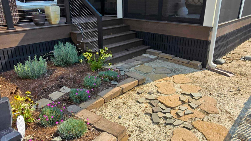 Backyard view of a screened-in porch with stairs, black railing, and outdoor furniture visible inside; stone pathway and gravel ground with potted plants nearby.