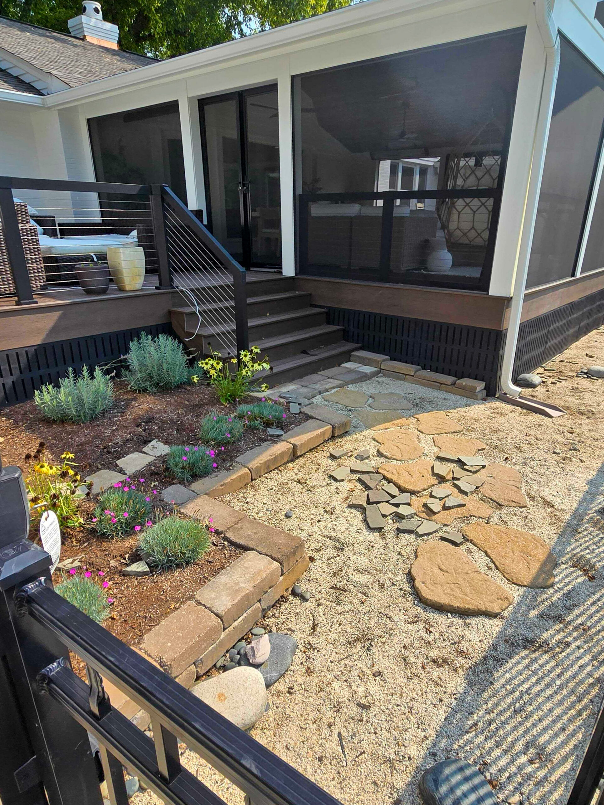Backyard view of a screened-in porch with stairs, black railing, and outdoor furniture visible inside; stone pathway and gravel ground with potted plants nearby.