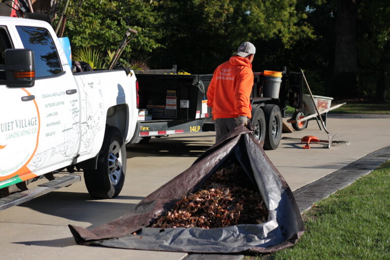 Lawn care worker in an orange hoodie stands beside a landscaping truck, unloading a tarp filled with leaves onto the curb.