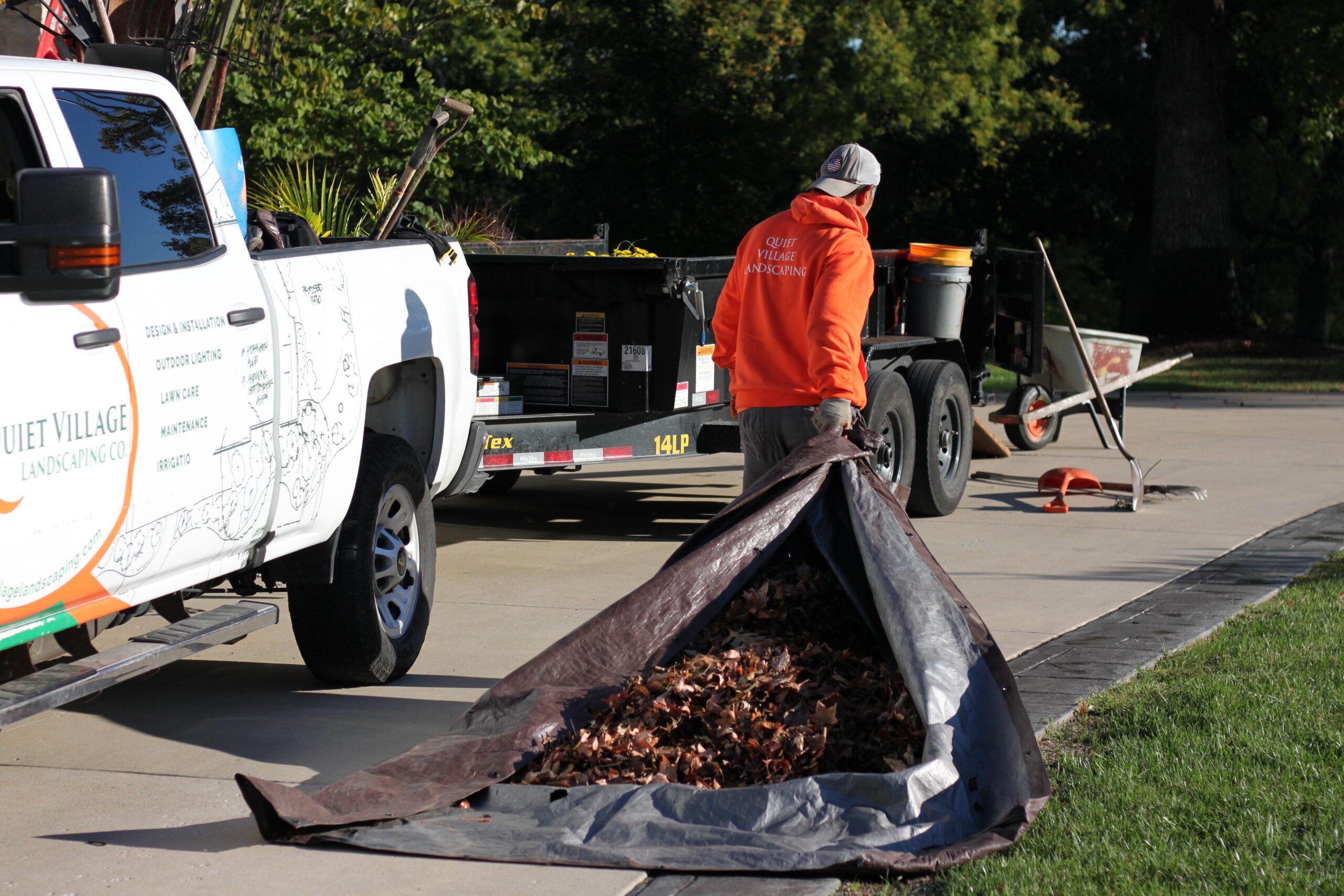 Lawn care worker in an orange hoodie stands beside a landscaping truck, unloading a tarp filled with leaves onto the curb.