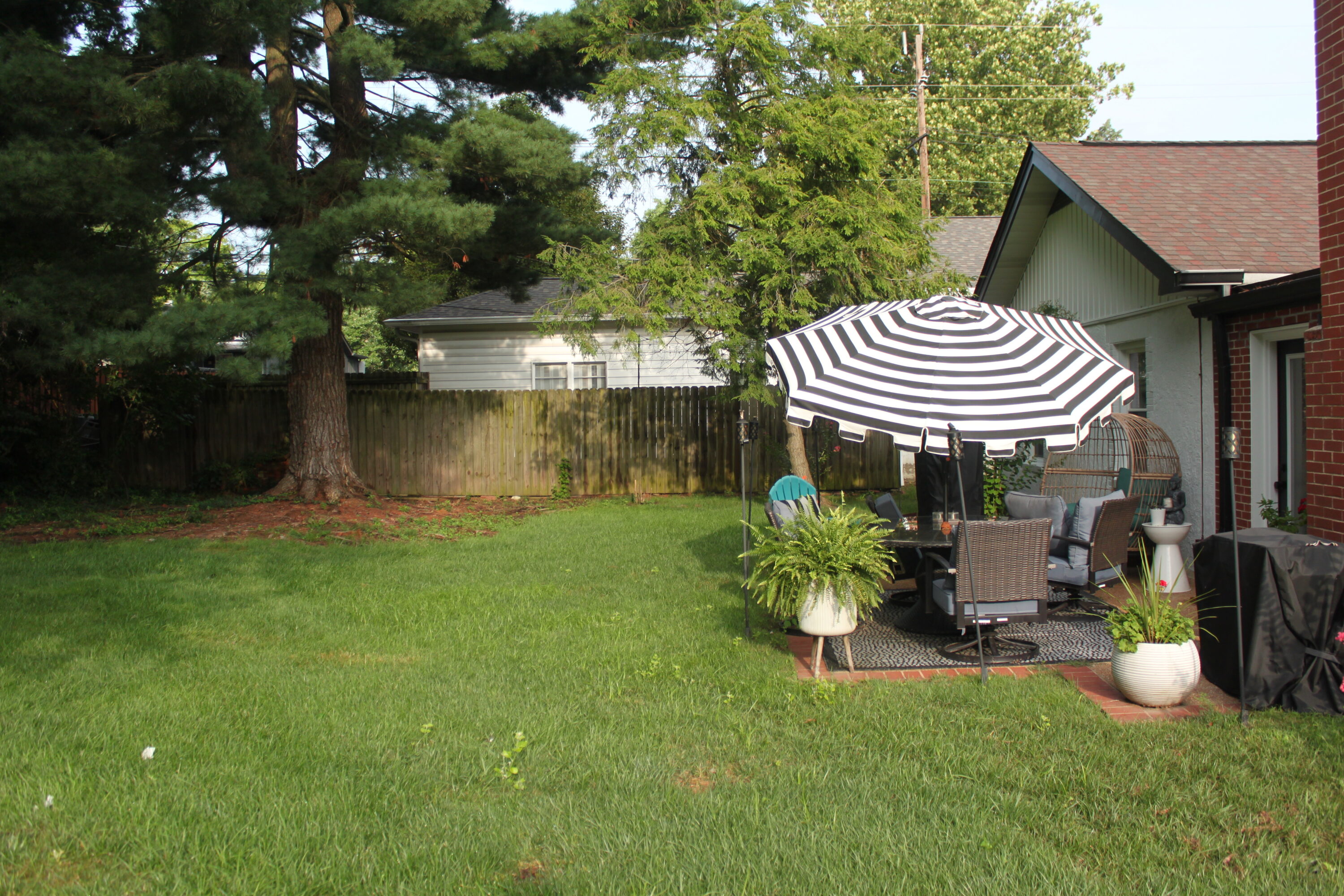 Backyard scene with a black-and-white striped patio umbrella over outdoor furniture on a brick-patio, green lawn and trees nearby.