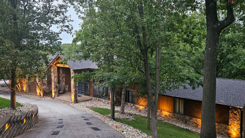 Tree-lined residential street with a stone-and-wood house and warm exterior lights.