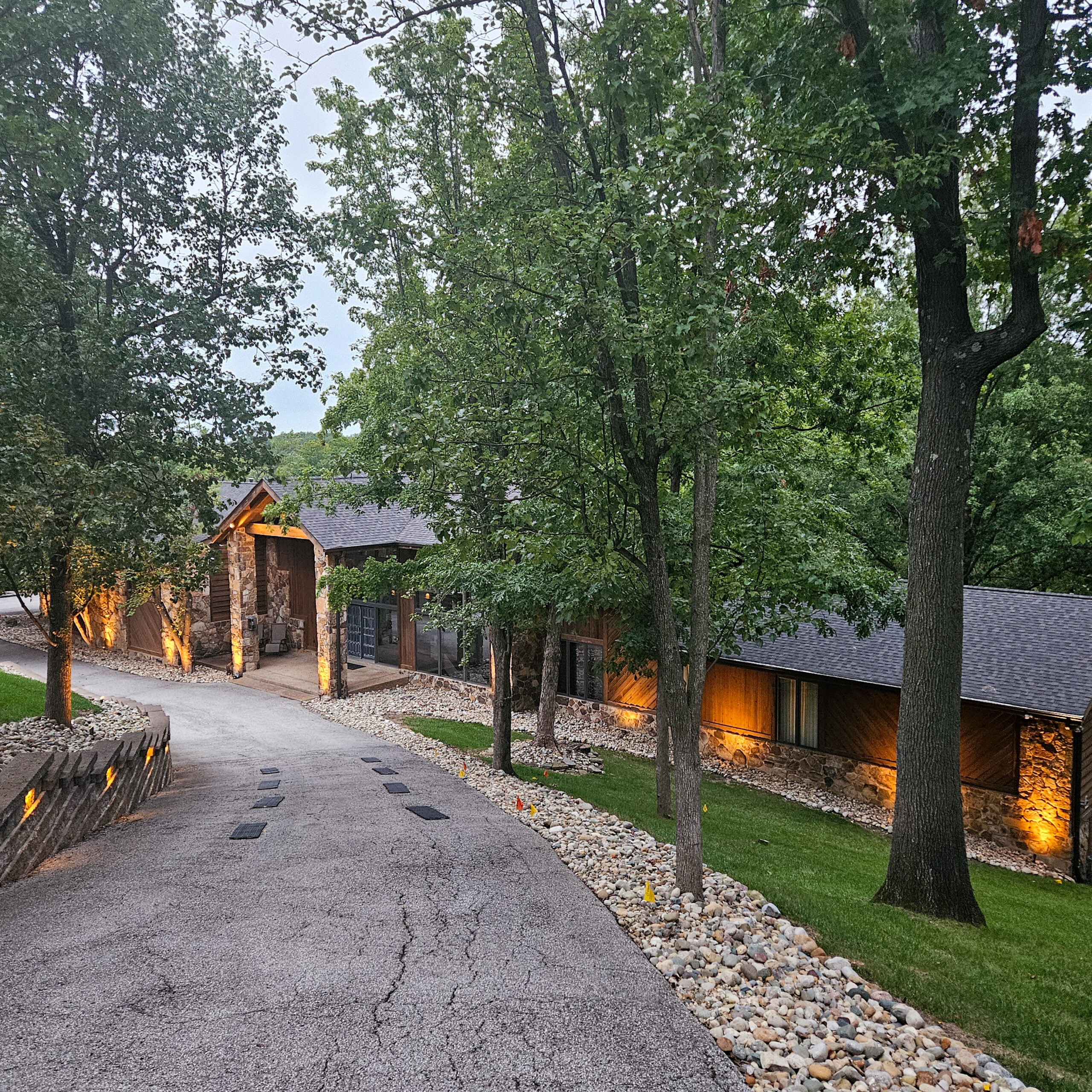 Tree-lined residential street with a stone-and-wood house and warm exterior lights.