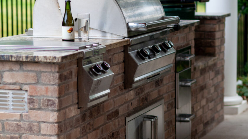 Outdoor kitchen with brick island grill, a bottle of wine and a glass on the counter in front of a stainless steel barbecue.