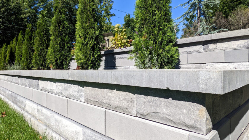 Tiered gray concrete retaining wall with evergreen trees and a bright blue sky in the background