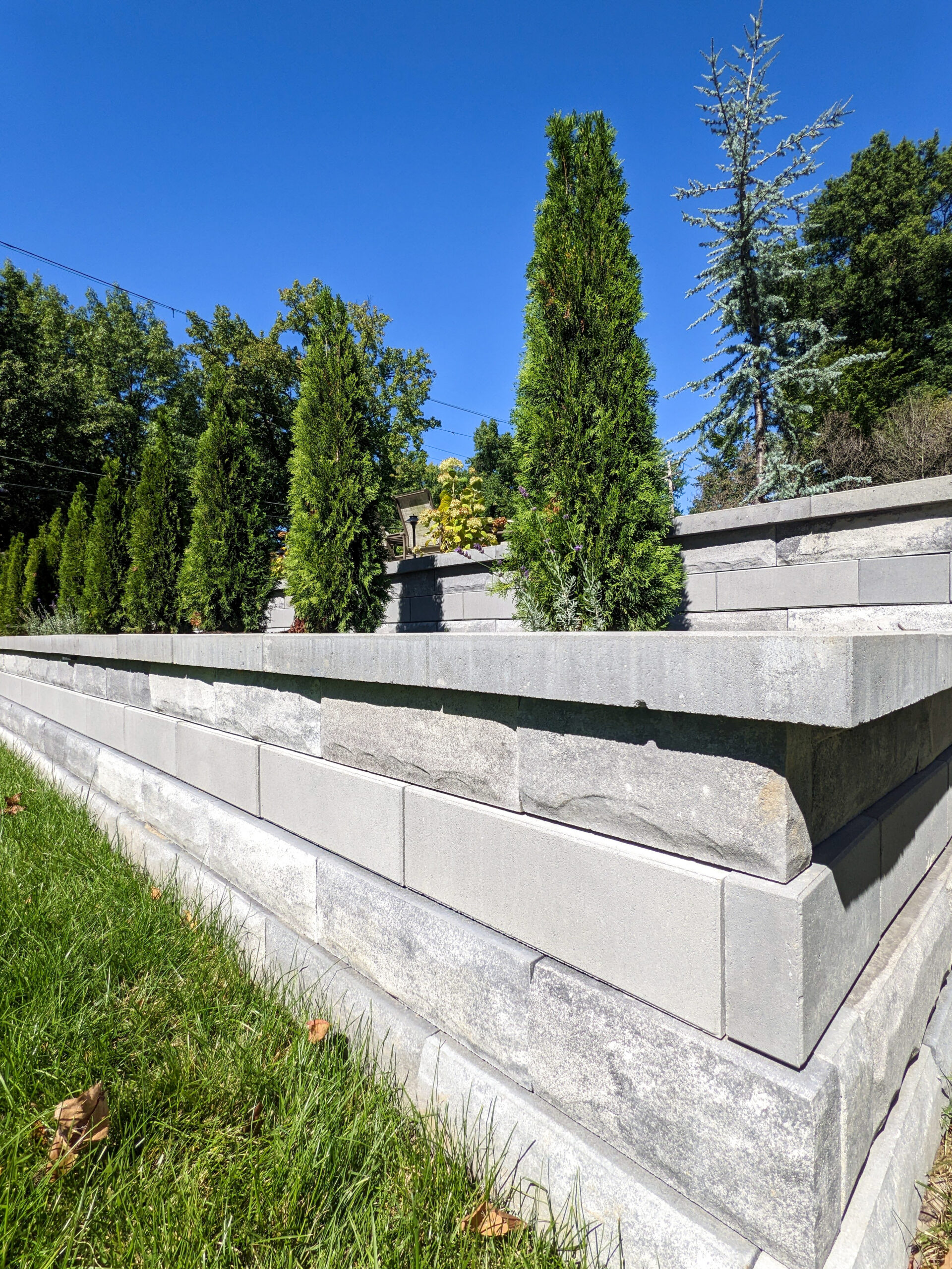 Clayton-RetainingWall14 | Quiet Village Landscaping Tiered gray concrete retaining wall with evergreen trees and a bright blue sky in the background