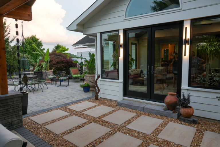 Patio area with beige stepping-stone pathway, gravel edging, and metal tables and chairs, surrounded by potted plants and outdoor lighting beside a modern house with large glass doors.