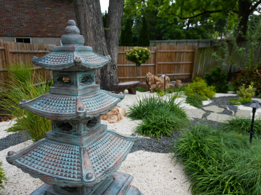 Close-up of a weathered, pagoda-style stone garden lantern in a gravel courtyard with plants and a wooden fence in the background.