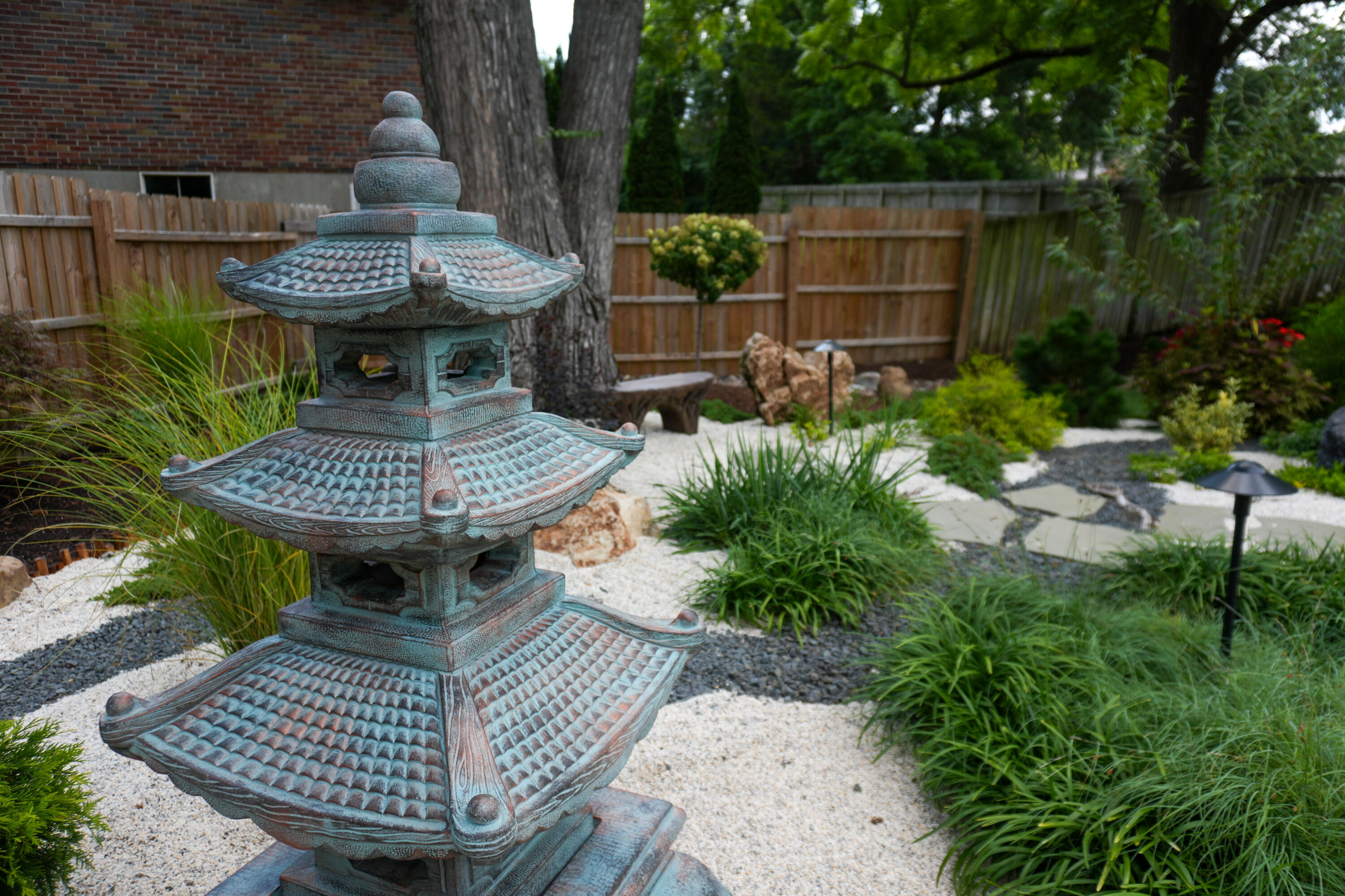 Close-up of a weathered Japanese stone lantern in a gravel garden, with a wooden fence and green trees in the background.