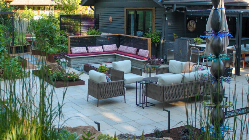 Cozy modern backyard patio with beige wicker seating, red cushions, and planter beds near a dark siding house.