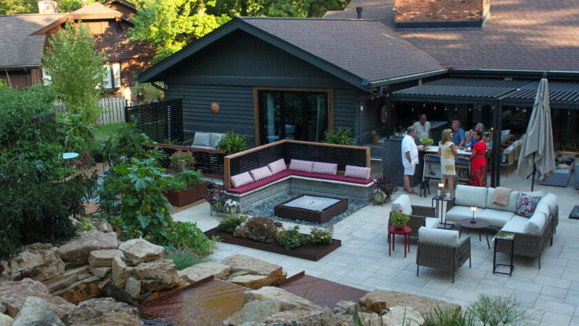 Backyard patio scene with dark siding house, outdoor seating, and a group of people chatting near a covered dining area; rock garden in the foreground.