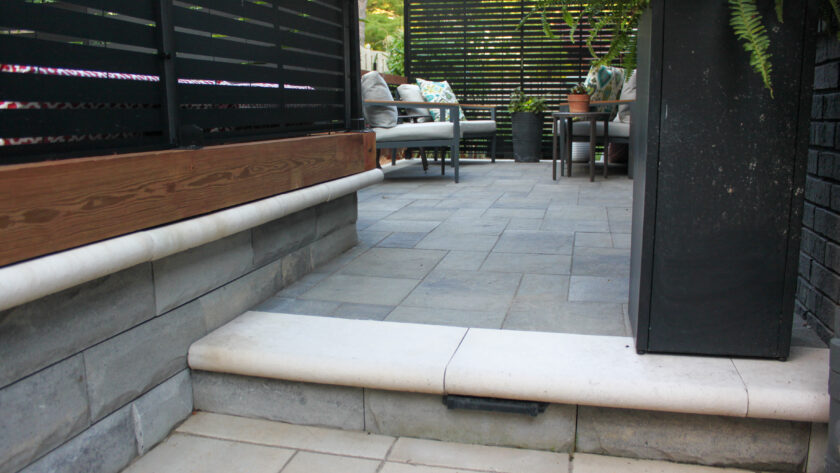 Outdoor patio scene with black slatted privacy walls, gray tile flooring, and white pipes along a raised concrete base; seating area visible in the distance.