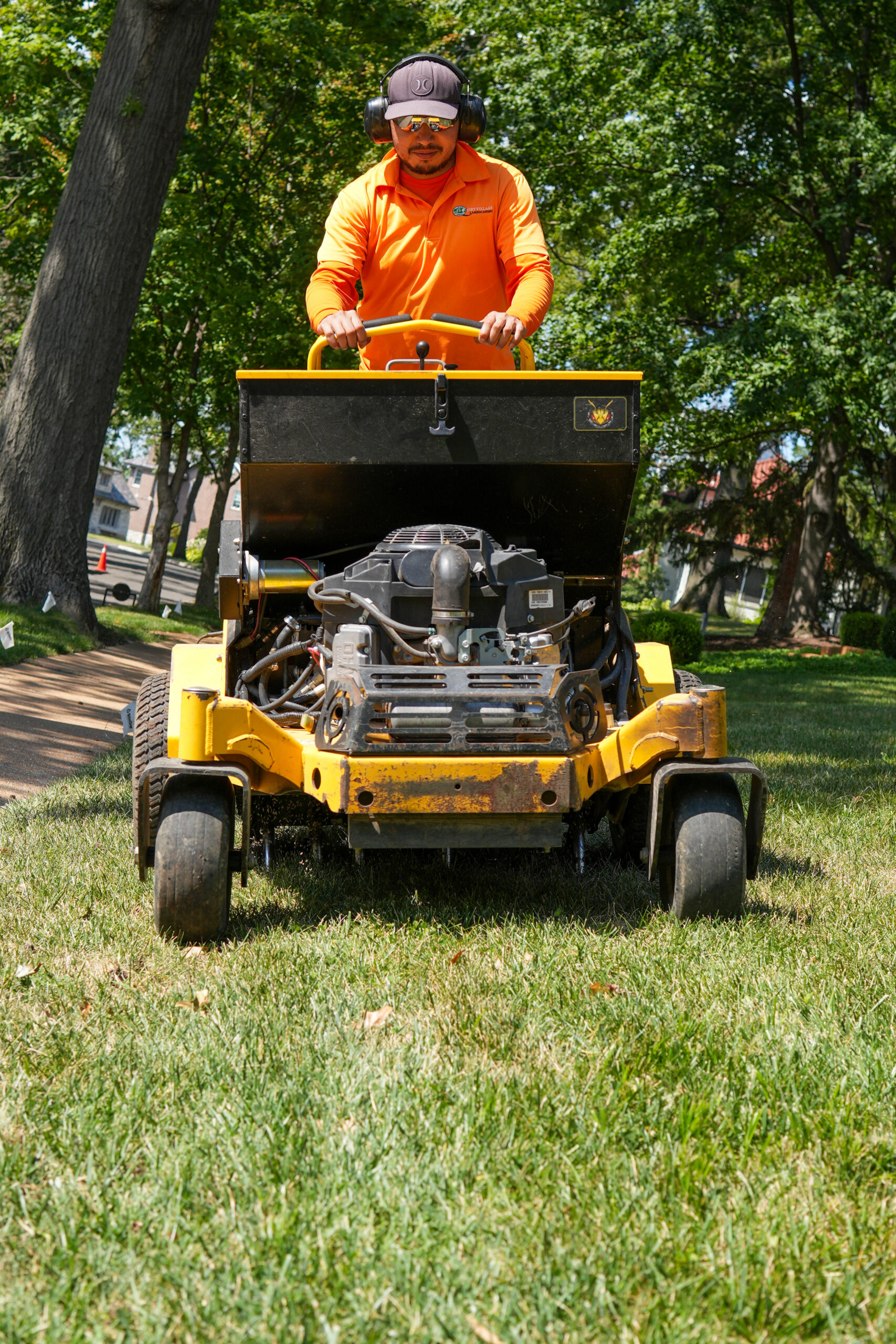 Man in an orange shirt operating a yellow riding lawn mower, wearing safety headphones and sunglasses, on a sunny residential lawn.