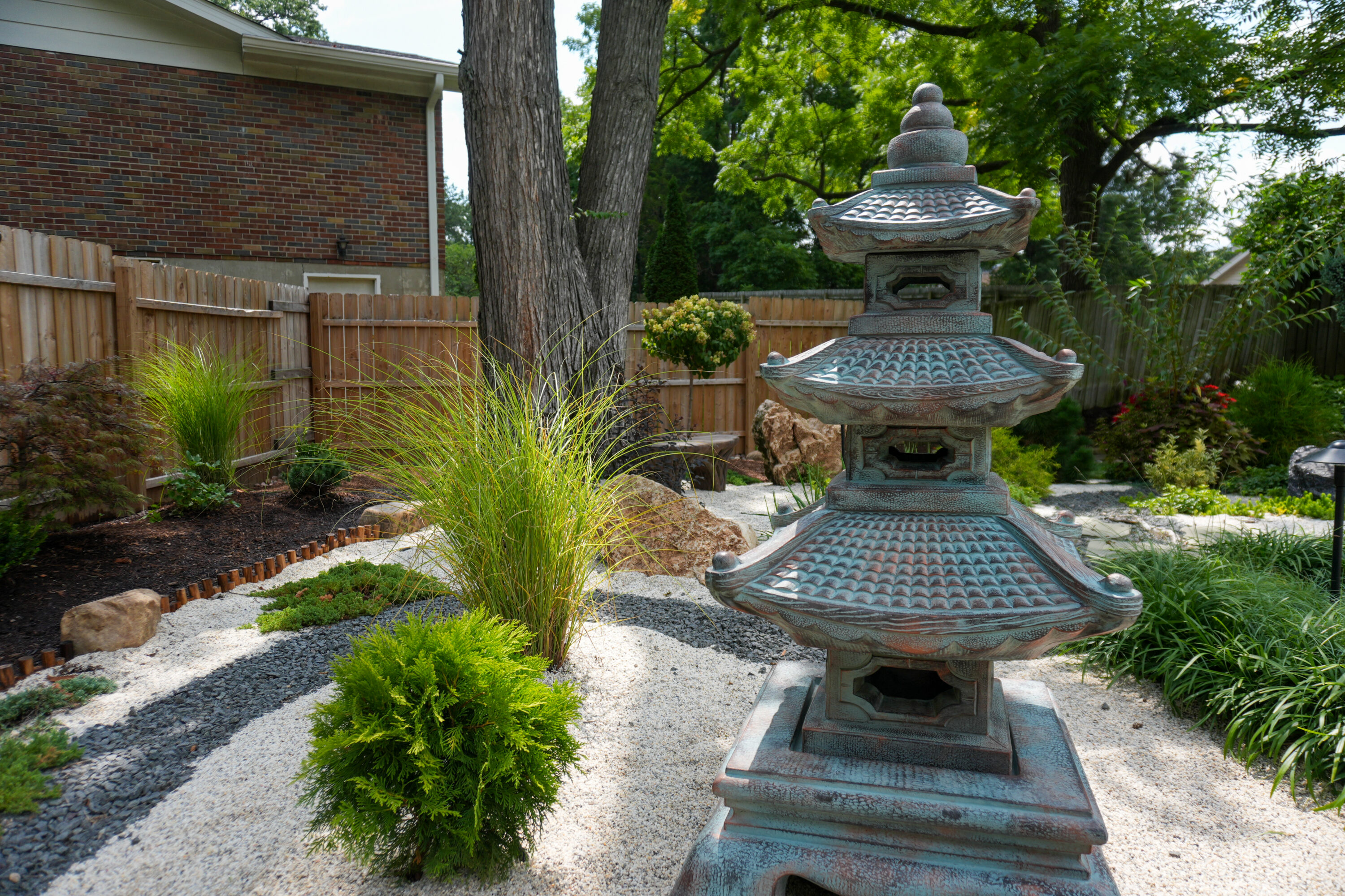 Tiered stone lantern in a Japanese-style garden, surrounded by gravel, grasses, and shrubs with a wooden fence and brick house in the background.