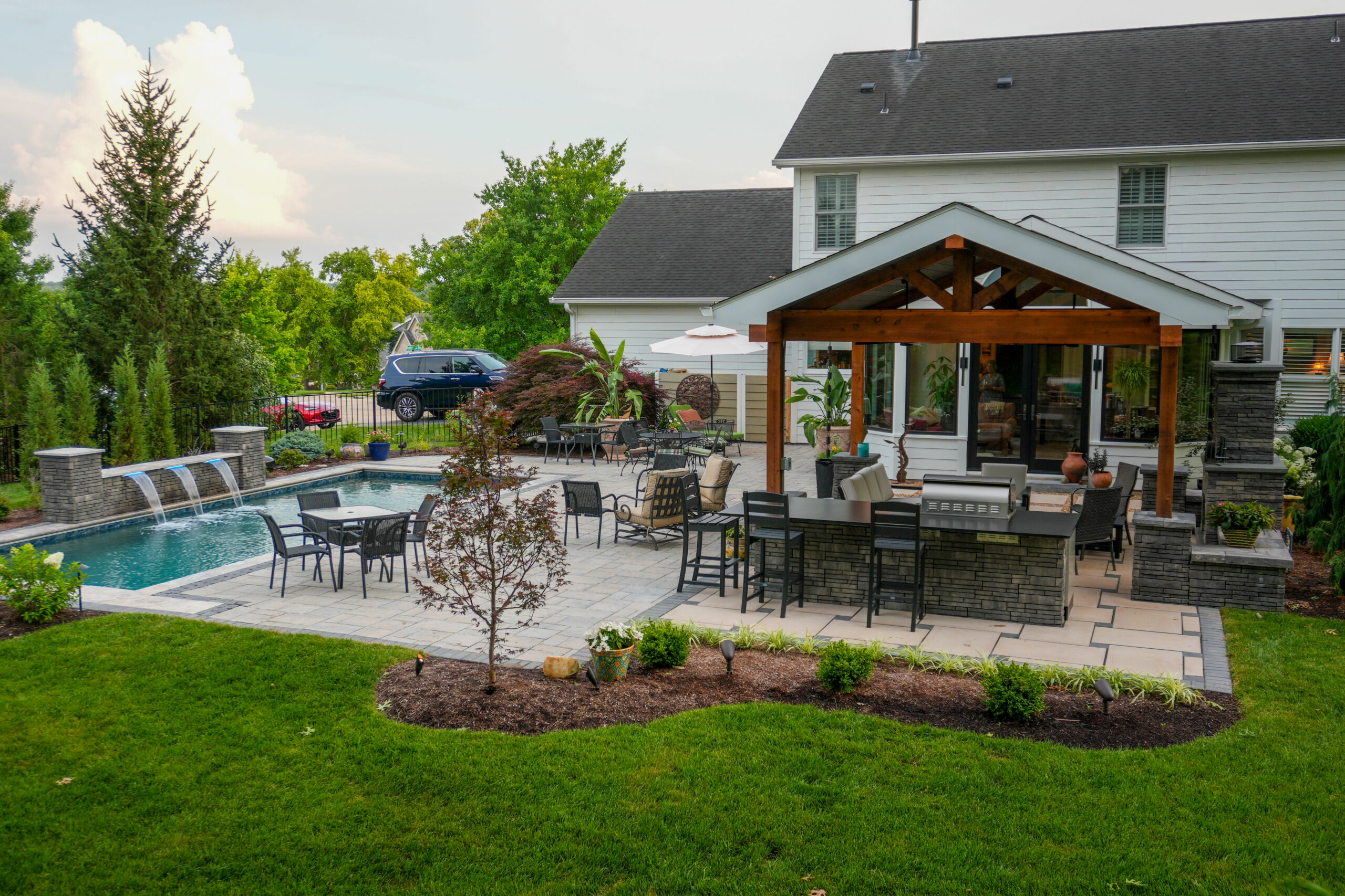 DesPeres-OutdoorKitchenPatio3 | Quiet Village Landscaping Backyard scene with an in-ground pool, stone patio, and seating under a wooden pergola beside a white house.