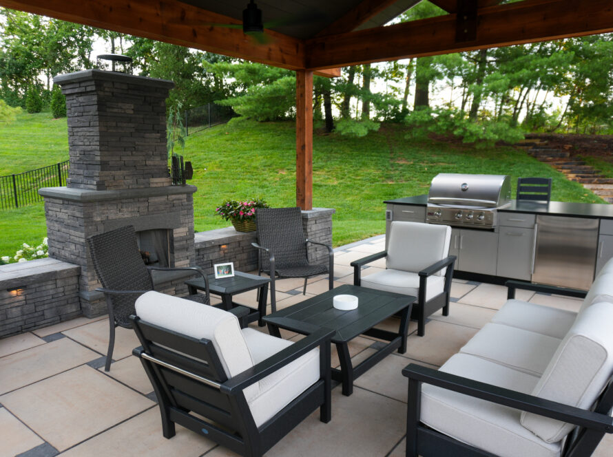 Outdoor patio with a stone fireplace, wicker chairs, and a stainless grill under a wooden cover overlooking a green lawn.