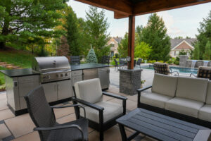 Outdoor patio with a stainless grill and counter, beige seating, and a view of a pool amid green trees and a house in the background.