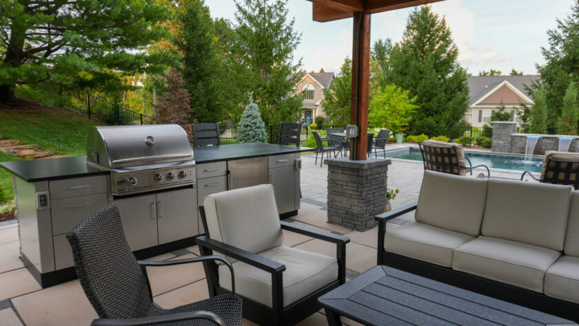 Outdoor patio with a stainless grill and counter, beige seating, and a view of a pool amid green trees and a house in the background.