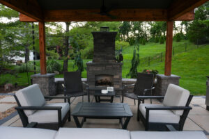 Outdoor seating area under a wooden patio cover with a stone fireplace and green lawn beyond.