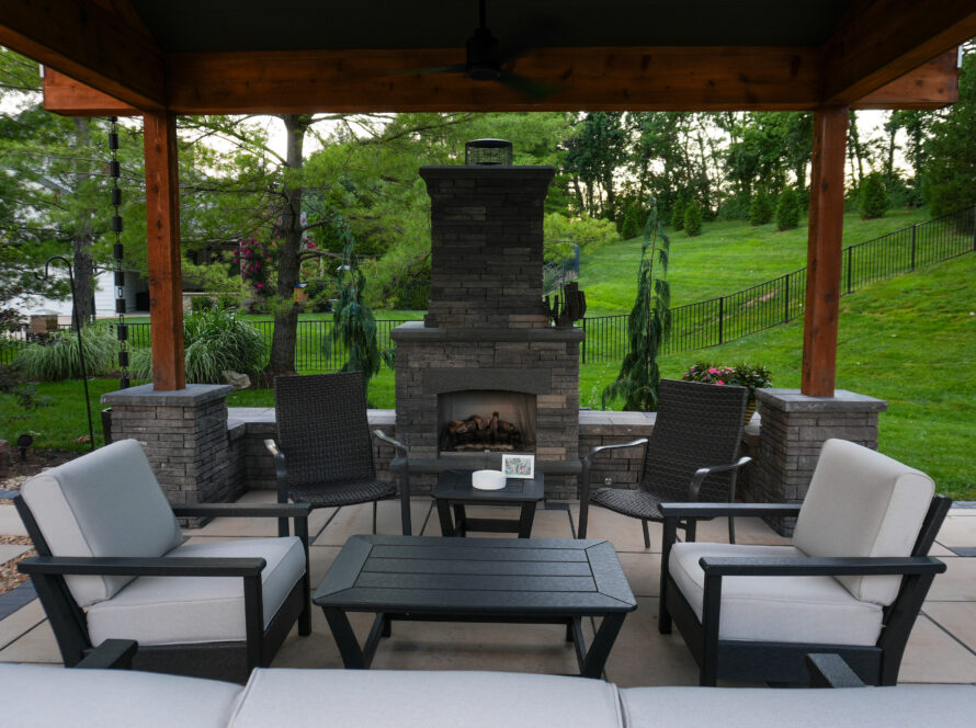 Outdoor seating area under a wooden patio cover with a stone fireplace and green lawn beyond.