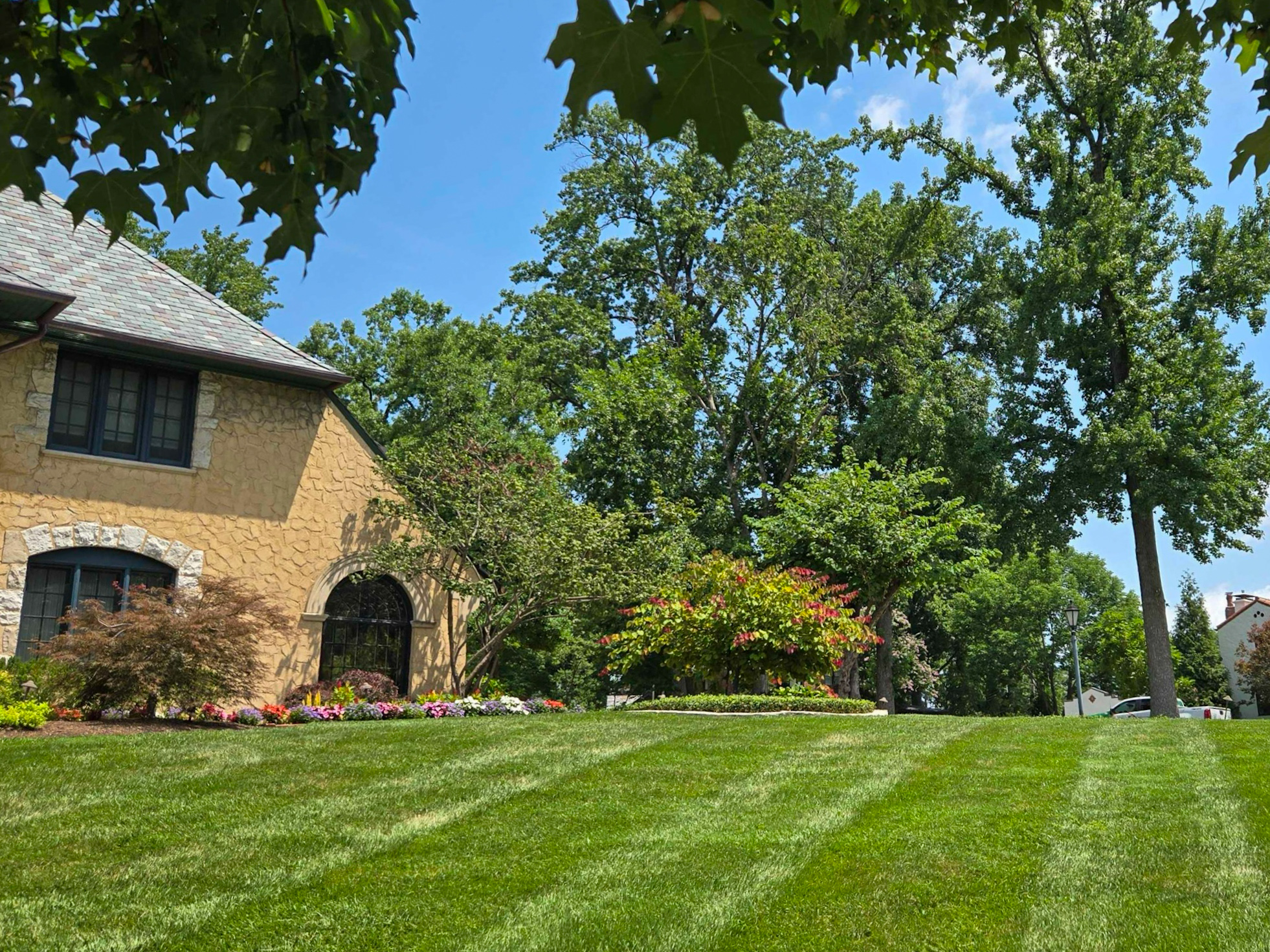 Sunlit front yard of a beige stone house with an arched window, surrounded by colorful flower beds and green lawn.