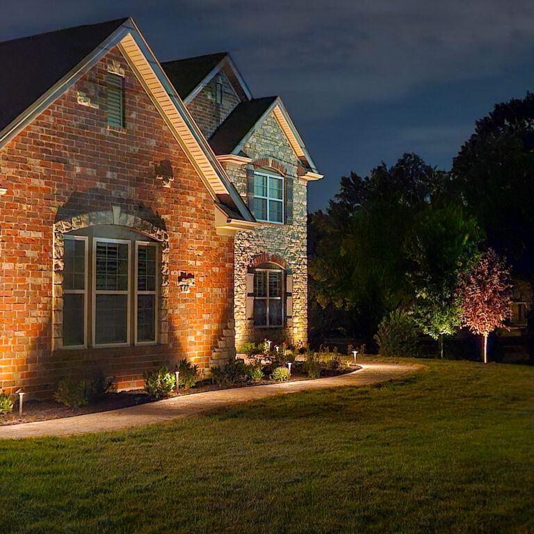Brick house at night with warm exterior lighting highlighting the brick walls and peaked rooflines, plus a well-kept lawn and landscaping.