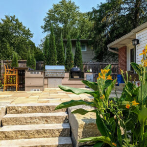 Backyard patio with stainless grill, charcoal cooker, and outdoor kitchen under a wooden fence with tall evergreens in the background.