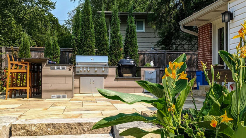 Backyard patio with stainless grill, charcoal cooker, and outdoor kitchen under a wooden fence with tall evergreens in the background.