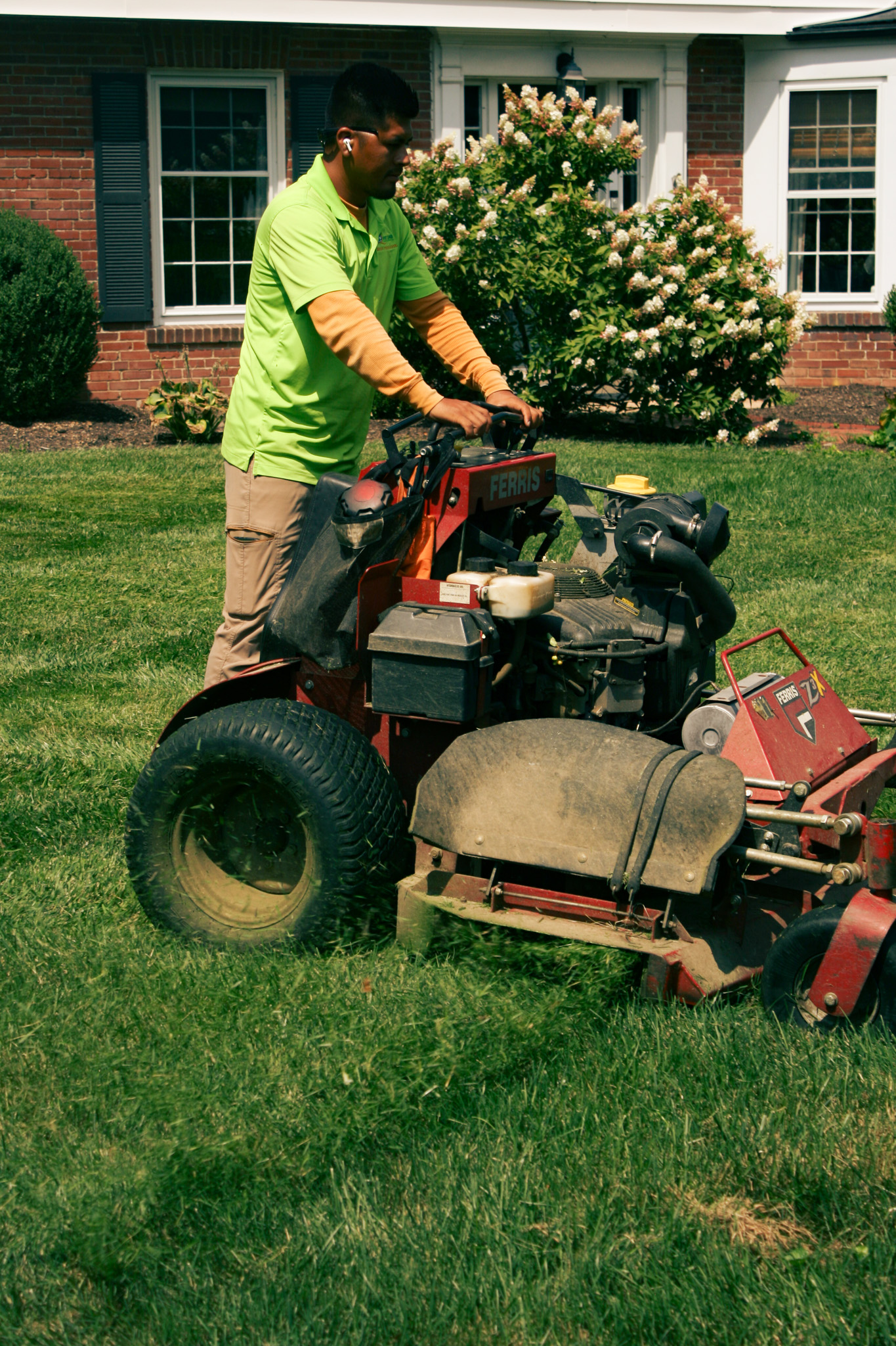 Man in a lime-green polo driving a red lawn mower on a front lawn with a flowering shrub in the background.