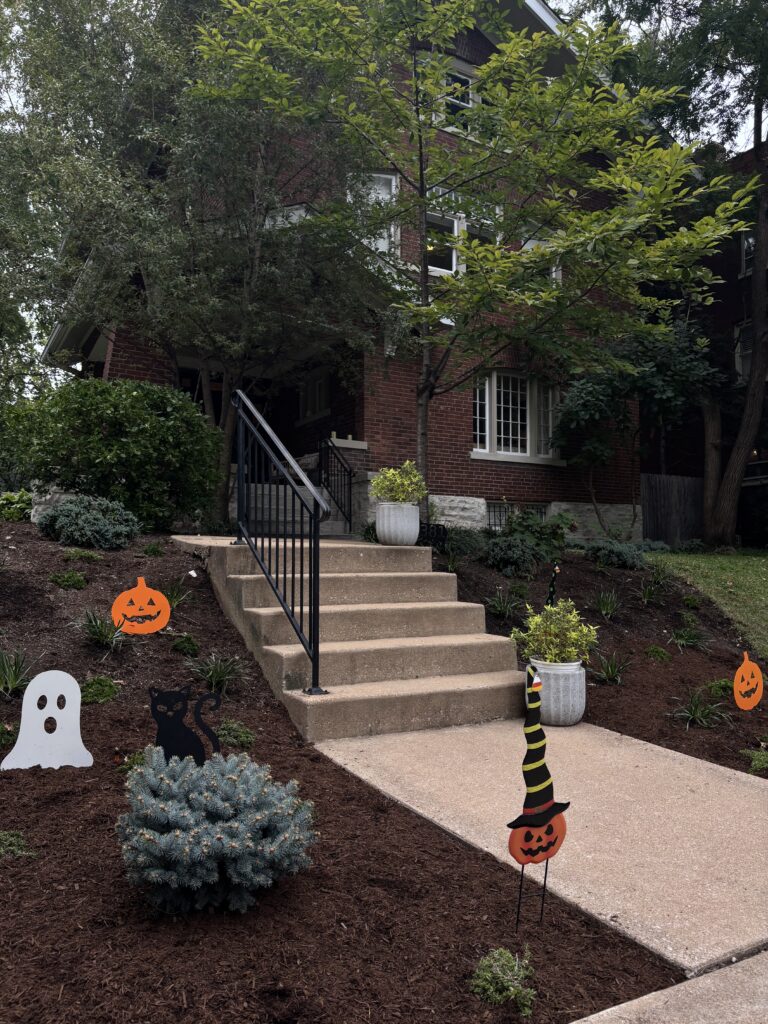 Brick house with front steps and porch, autumn trees, and Halloween decorations including jack-o'-lanterns, ghosts, and a black cat cutout kidney cake? wait no