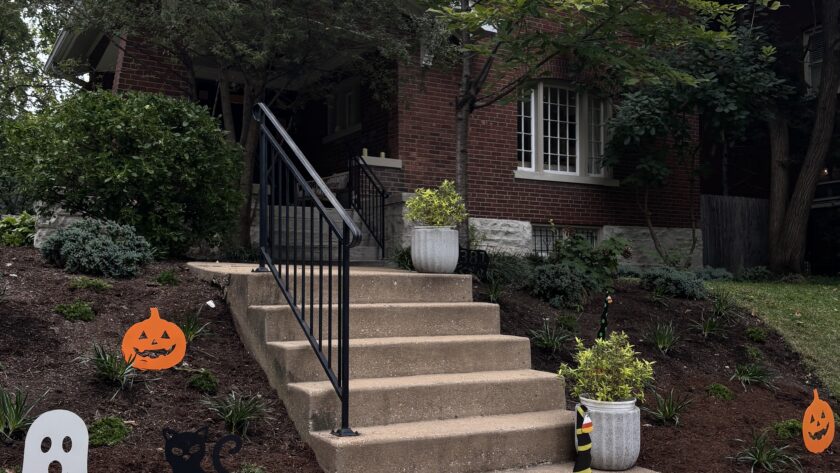 Brick house with front steps and porch, autumn trees, and Halloween decorations including jack-o'-lanterns, ghosts, and a black cat cutout kidney cake? wait no
