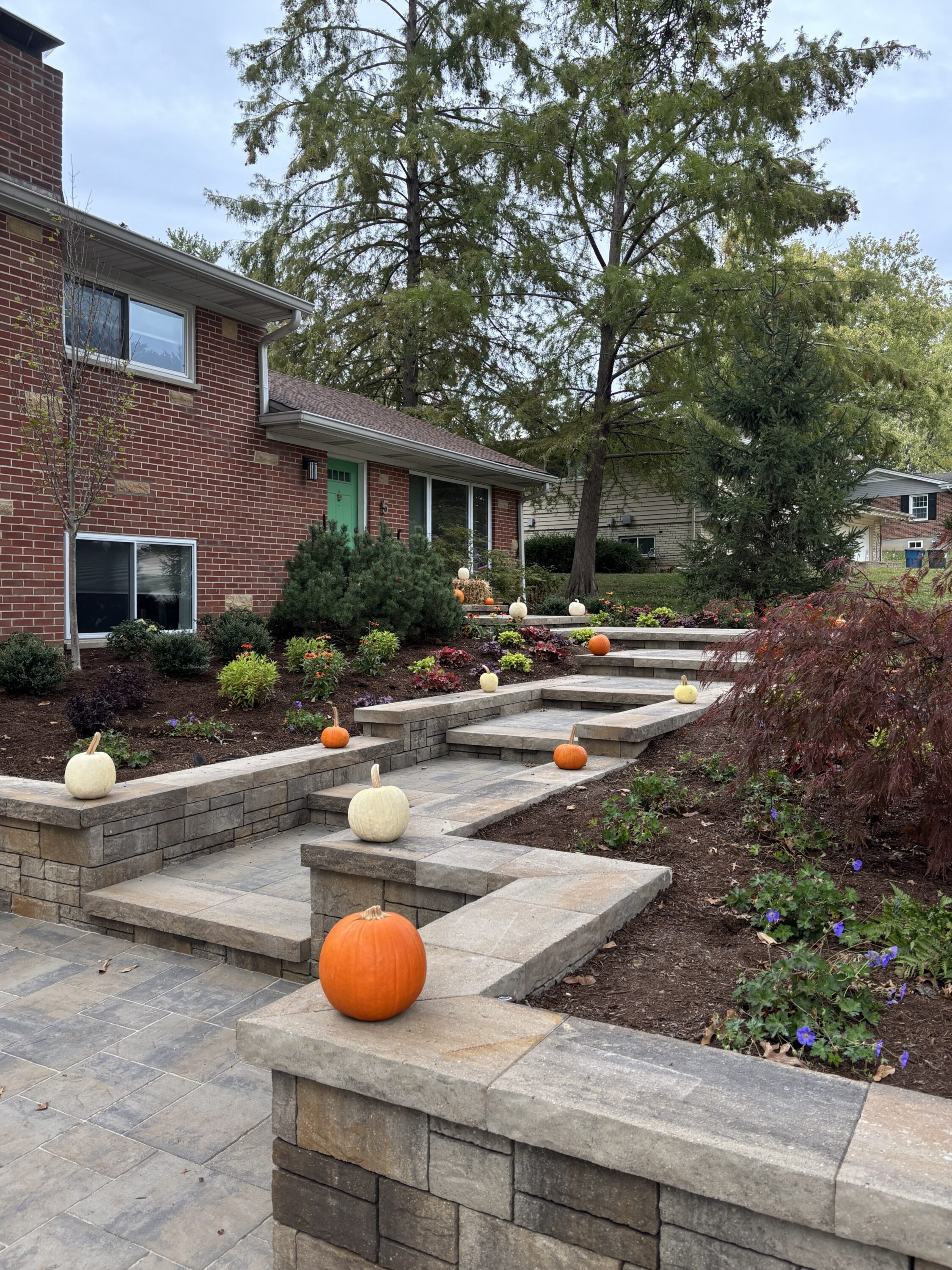 Brick house with a green front door, stone stepped planters, and pumpkins scattered along the front steps for autumn decor.