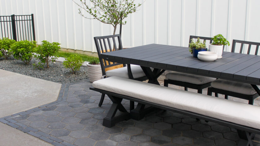 Outdoor black dining table with attached benches on hexagonal paving, several potted plants on and around the table, against a white wall backdrop.