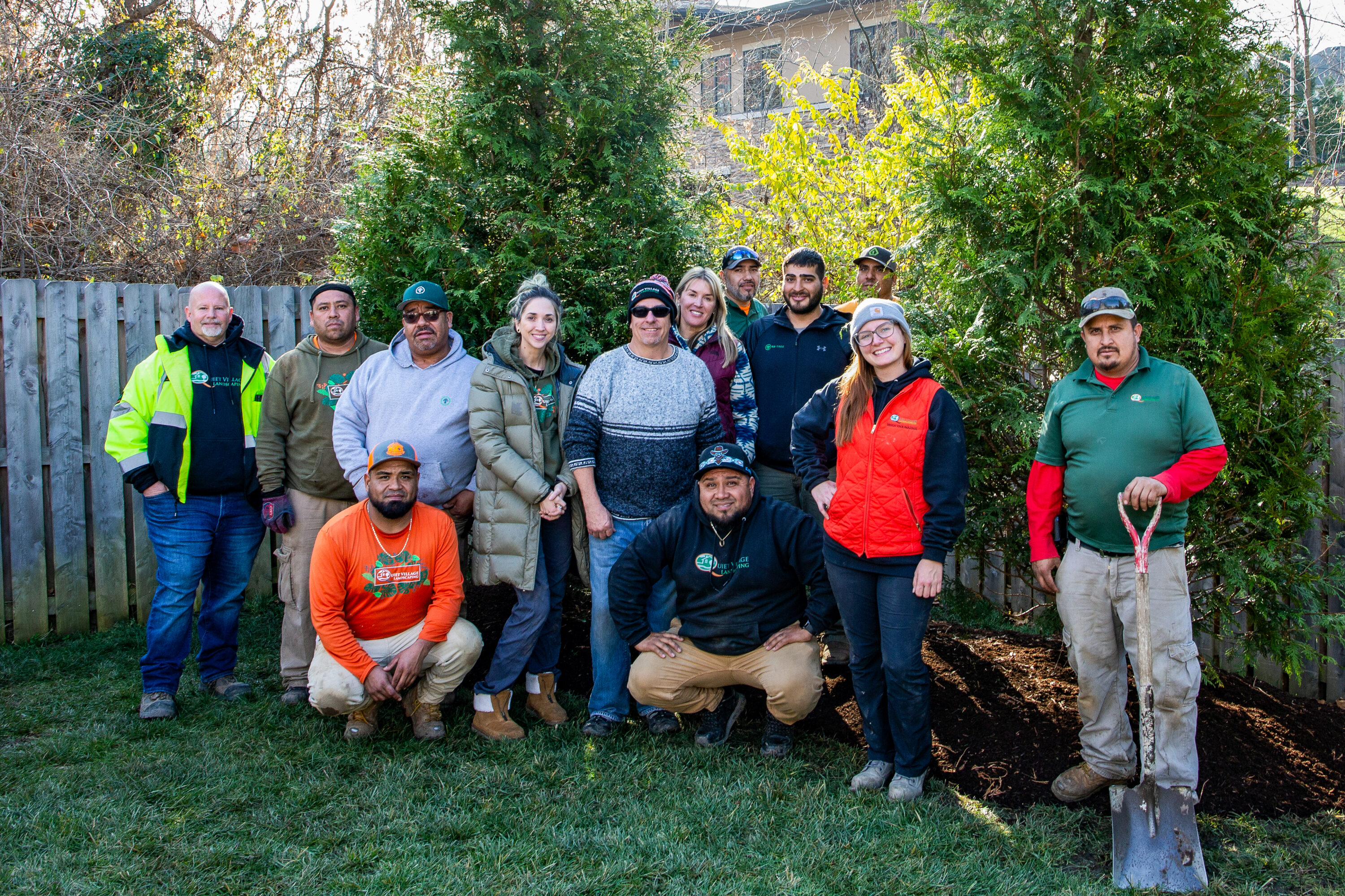 Group of diverse adults posing together outdoors in a backyard garden with trees and a wooden fence behind them.