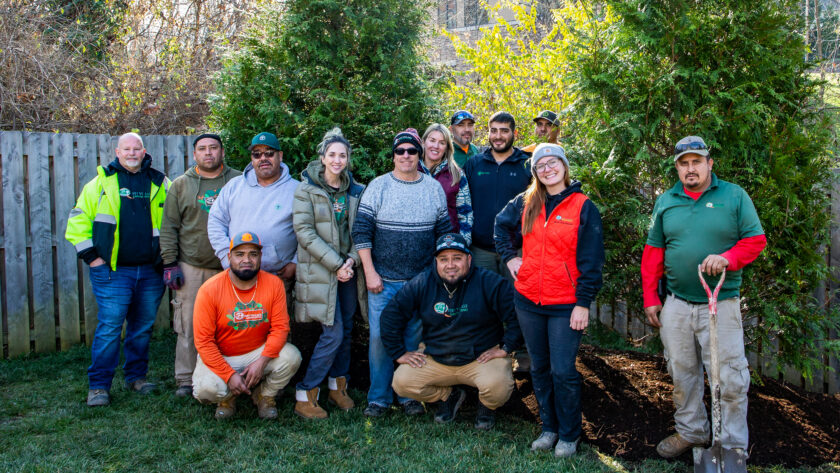 Group of diverse adults posing together outdoors in a backyard garden with trees and a wooden fence behind them.