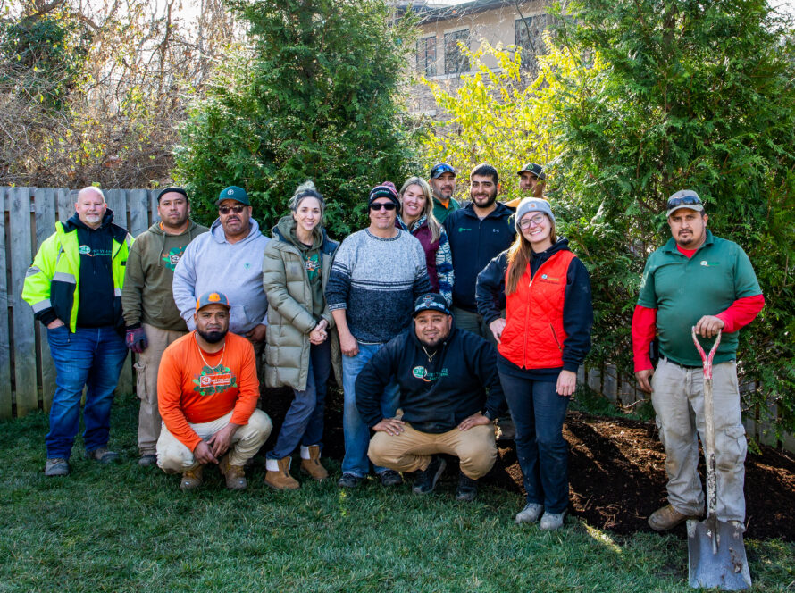 Group of diverse adults posing together outdoors in a backyard garden with trees and a wooden fence behind them.
