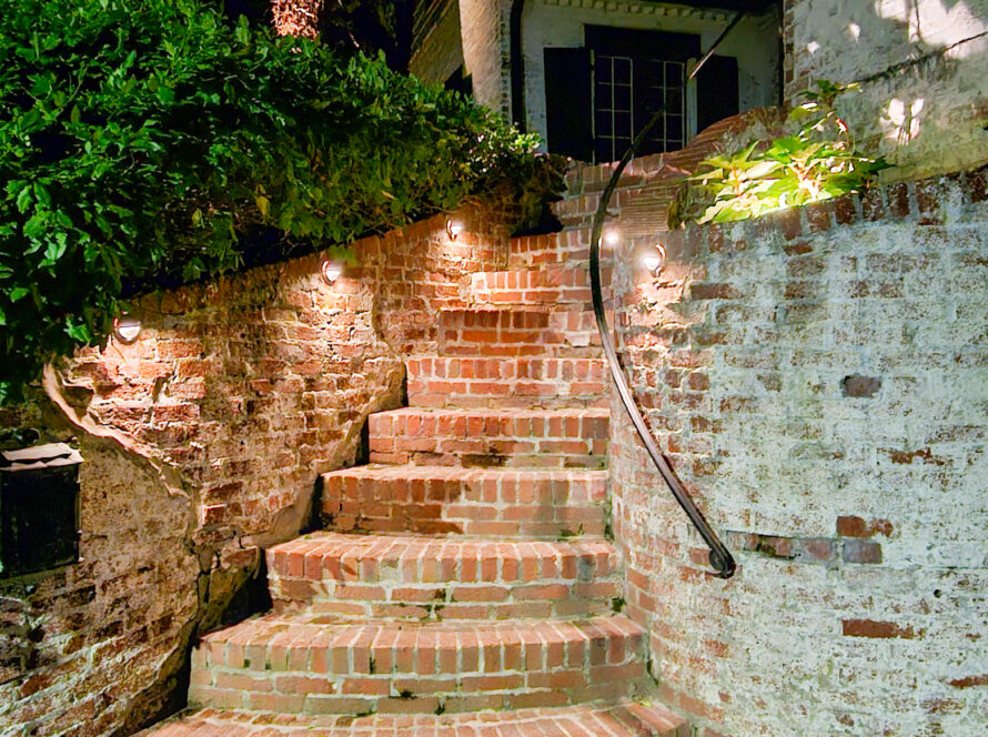 Nighttime brick staircase winding up a stone wall, ivy over a left hedge, with warm wall lights along the brick wall.