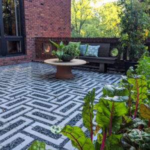 Brick-walled outdoor patio with a geometric white-and-gray patterned floor, round clay table, and a bench with blue-green cushions surrounded by greenery.