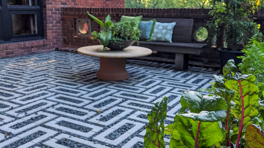 Brick-walled outdoor patio with a geometric white-and-gray patterned floor, round clay table, and a bench with blue-green cushions surrounded by greenery.