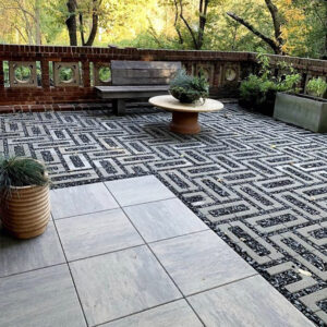 Outdoor patio with a black-and-white maze-patterned ground, a round planter-topped table, bench, and potted plants along a brick railing. The scene is sunny with trees in the background.