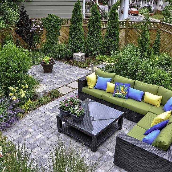 Outdoor patio with green cushioned wicker sectional, colorful pillows, a glass coffee table, and a surrounding garden.