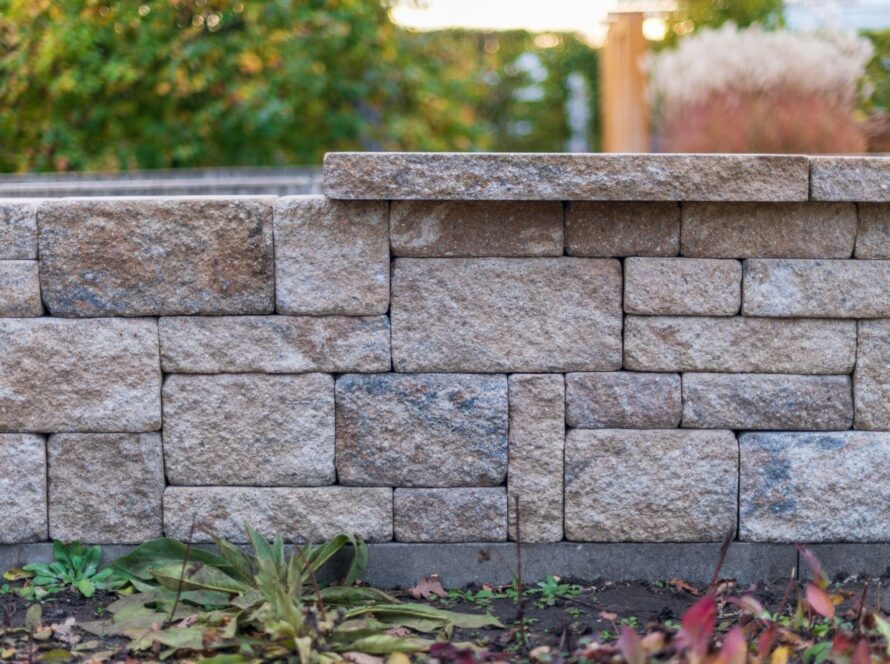 Stacked stone wall made of rectangular beige-gray blocks with green plants in the foreground and a blurred garden behind.