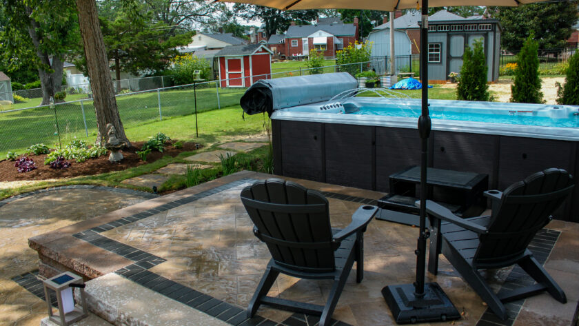 Backyard scene with a rectangular pool under a large umbrella, surrounded by patio chairs and a stone-tile patio.