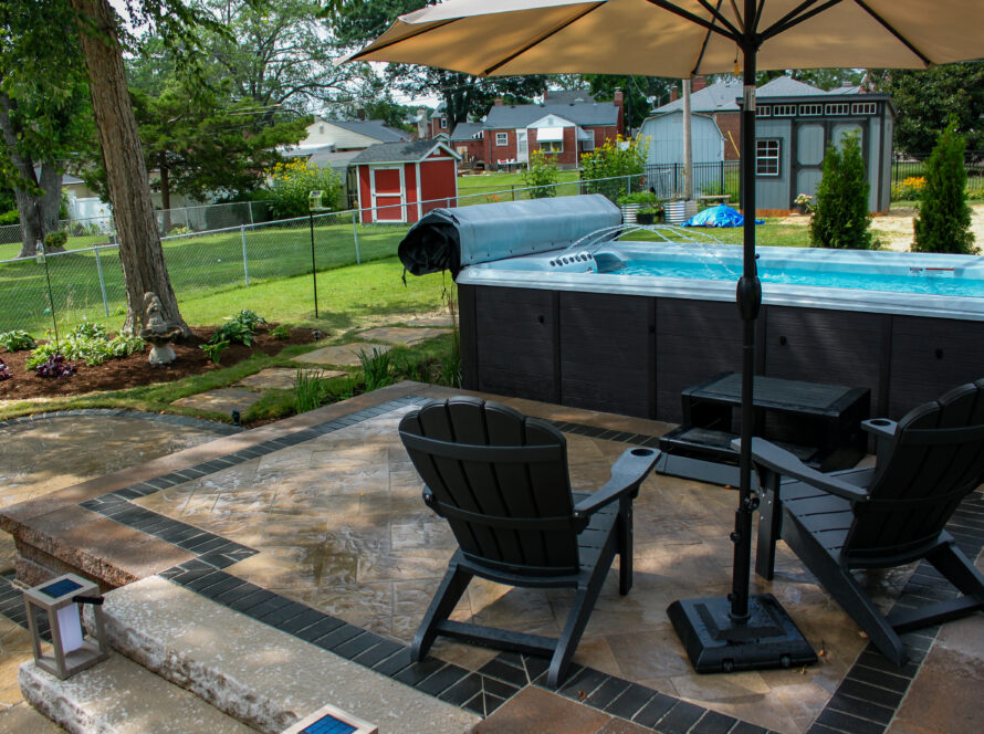 Backyard scene with a rectangular pool under a large umbrella, surrounded by patio chairs and a stone-tile patio.