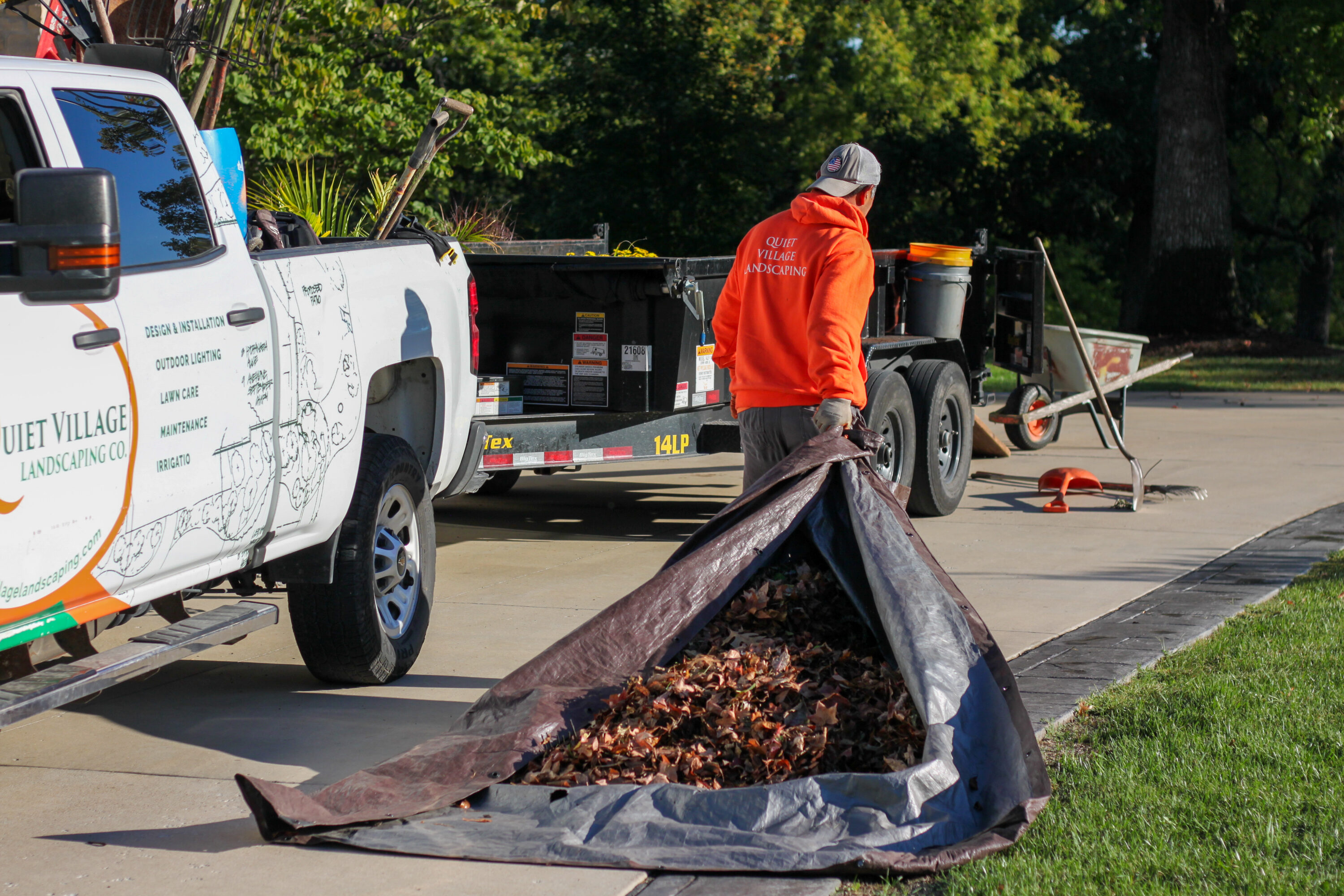 Man in orange hoodie standing beside a landscaping truck and trailer, loading a tarp with fallen leaves into a yard cleanup.