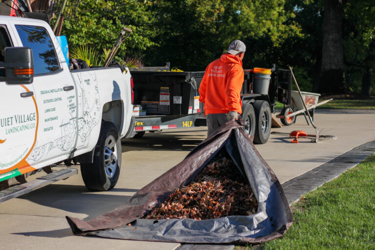 Man in orange hoodie standing beside a landscaping truck and trailer, loading a tarp with fallen leaves into a yard cleanup.