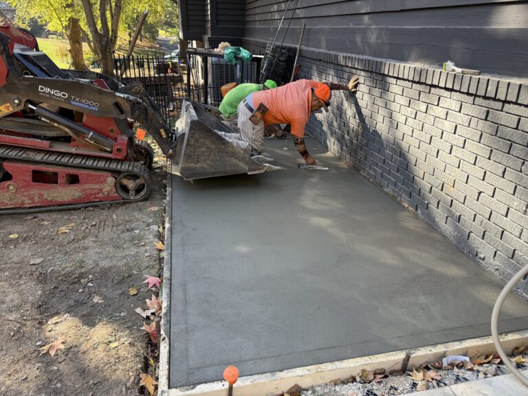 Construction worker in an orange shirt and bright cap smoothing freshly poured concrete along a house foundation, with a red skid-steer loader nearby.