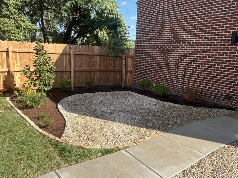 Curved gravel garden bed along a brick house wall, with small shrubs, a wooden fence, and trees in the background on a sunny day