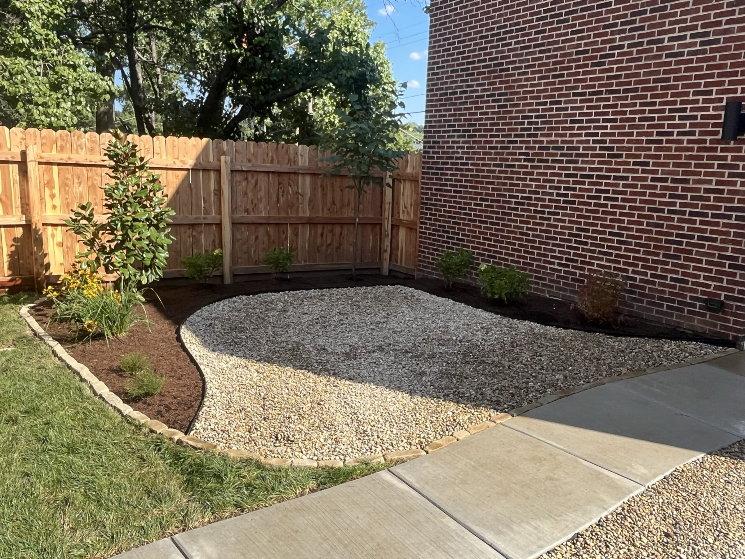 Curved gravel garden bed along a brick house wall, with small shrubs, a wooden fence, and trees in the background on a sunny day