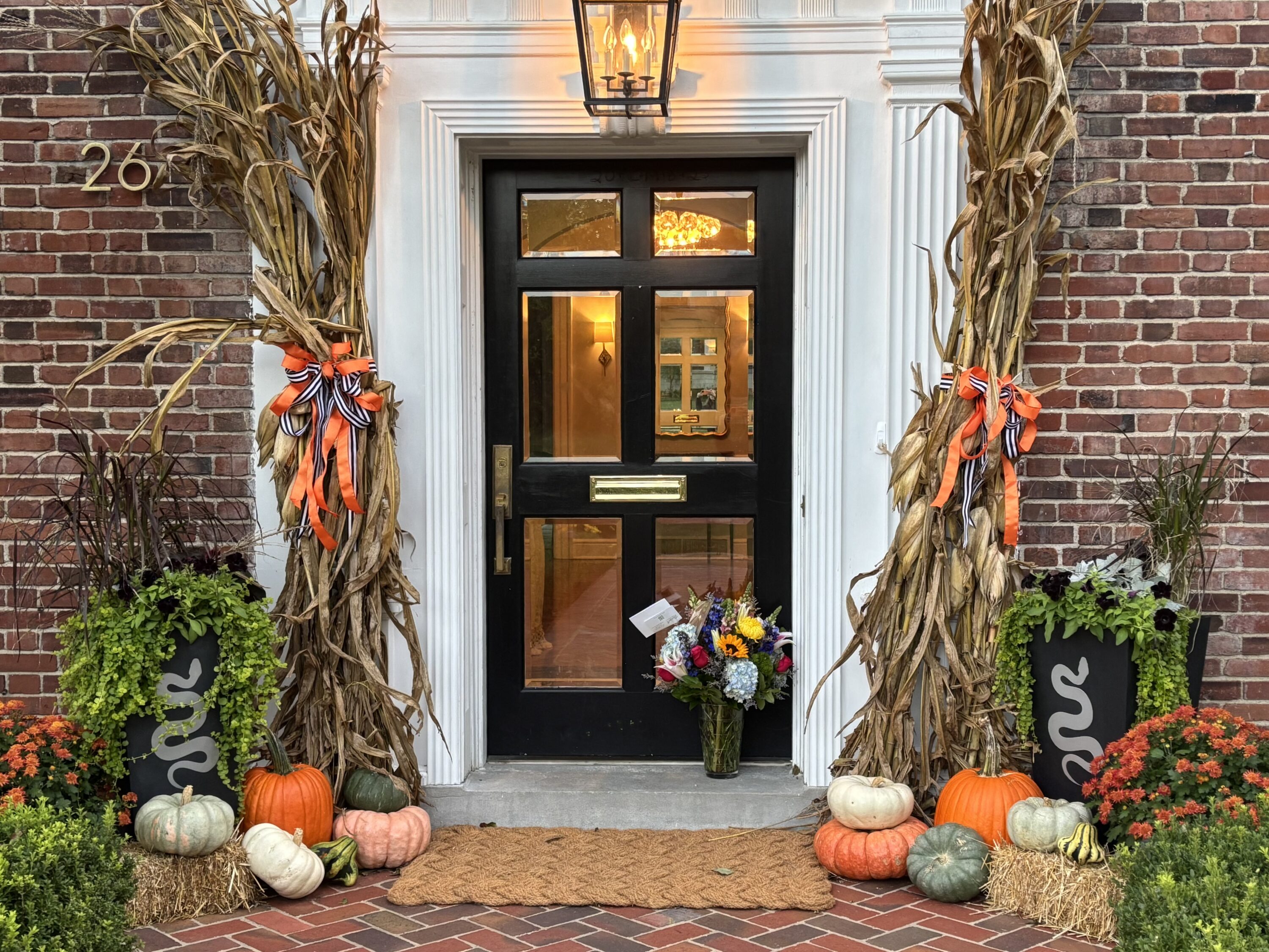 Front door framed by white trim and brick, with harvest decor including cornstalks, pumpkins, and mums on the steps.